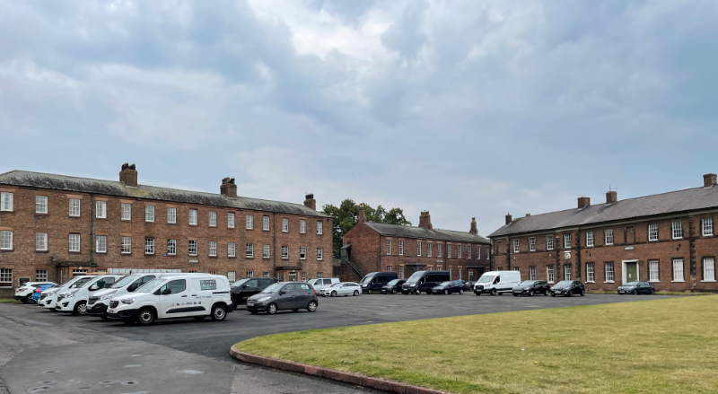 Long rectangular brick buildings with rows of sash windows. An impressive array of white vans is parked in front of them.
