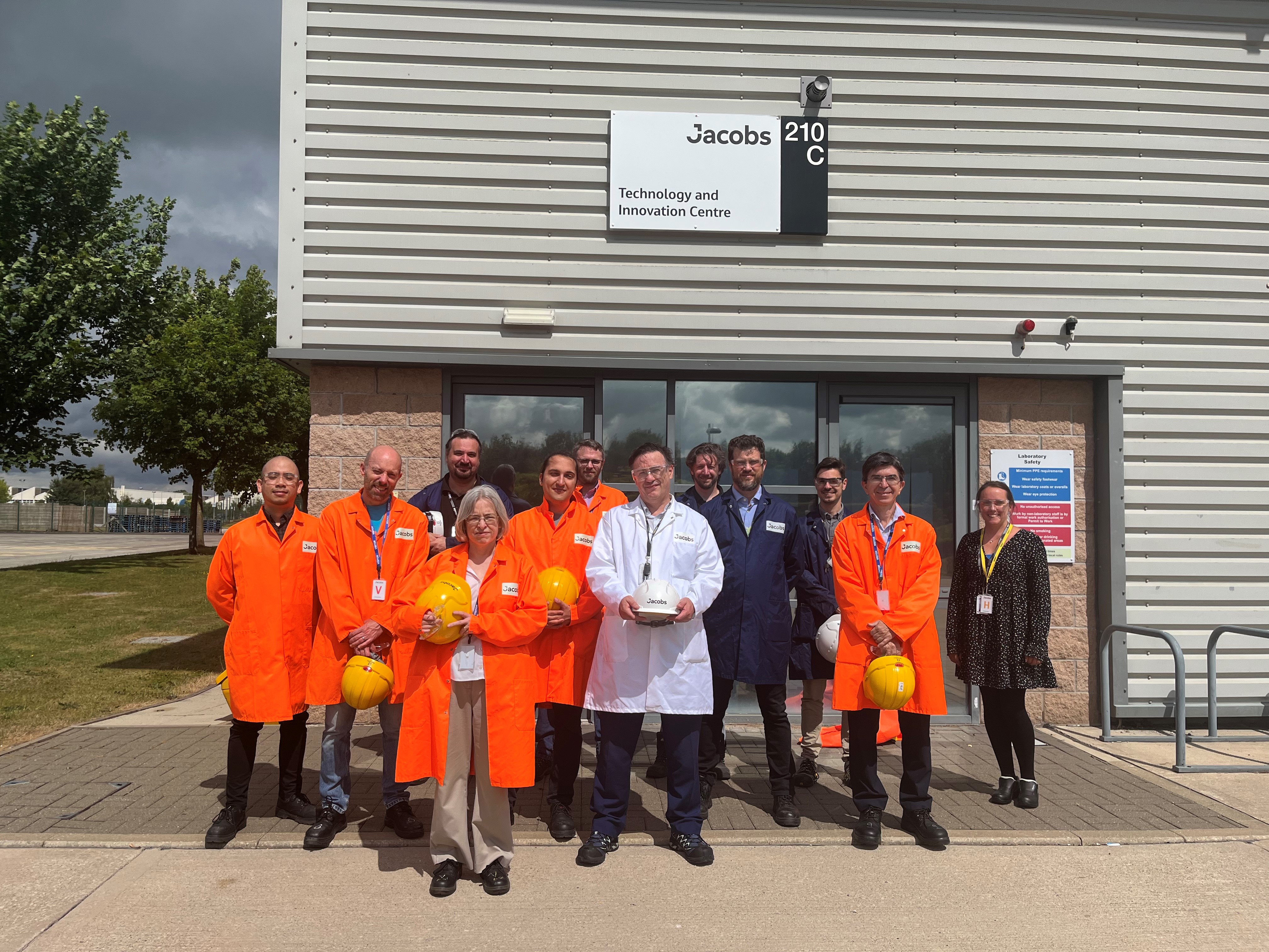A group of people in overalls holding hard hats, squinting into the sun. Most of the overalls are bright orange. I am at the front. We are standing in front of a building signed Jacobs 210 C Technology and Innovation Centre.